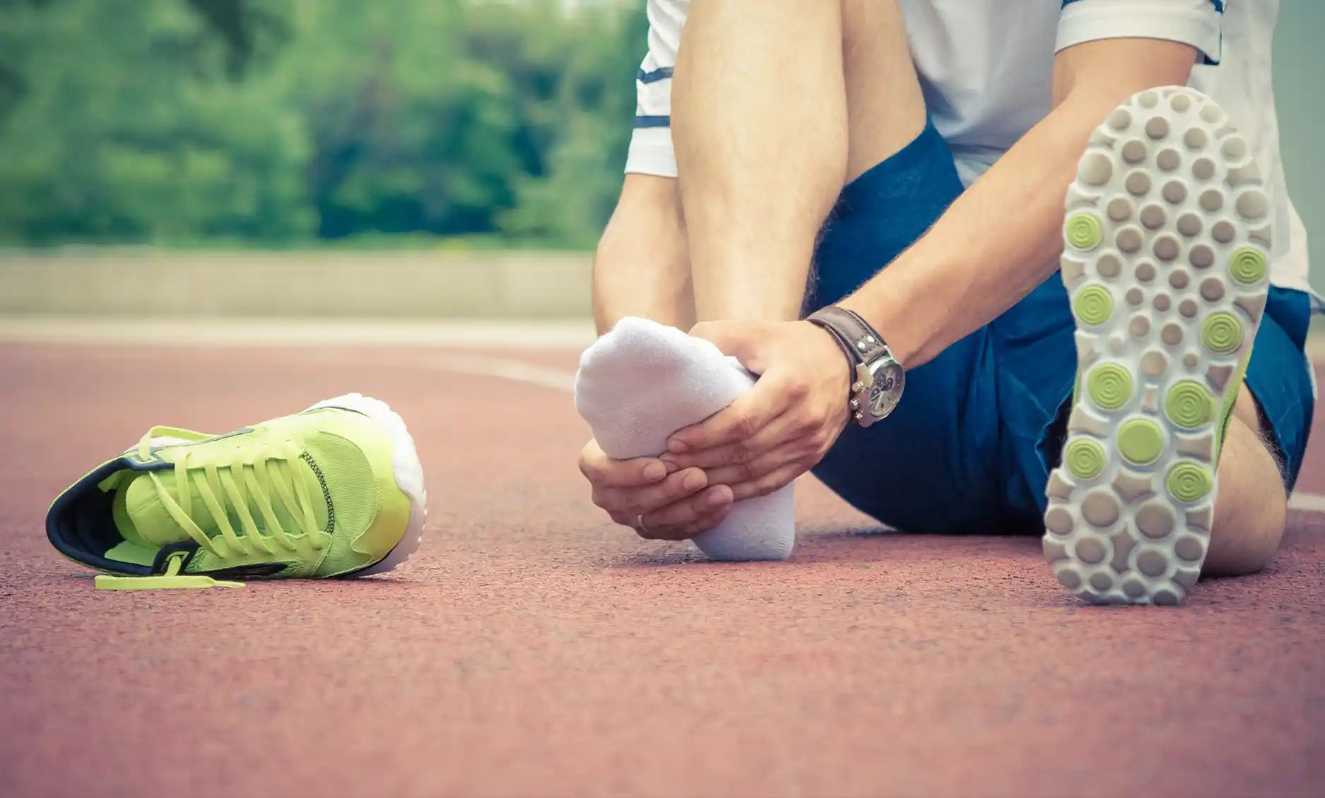 A person seated on a track holds their foot, indicating discomfort. Visit Foot & Ankle Center of Arizona for expert care.